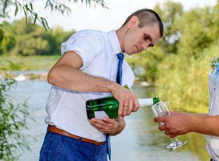 Bridegroom pouring a bottle of champagne into two flutes held by a wedding guest or best man as they celebrate at the wedding reception alongside a riverの写真素材