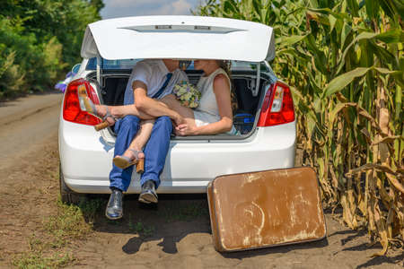 Unidentifiable married couple obscured by car trunk outside beside cornfield. Large brown suitcase on ground.の写真素材