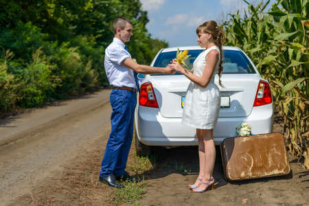 Young man giving a woman an ear of corn near rear of white automobile near cornfieldの写真素材