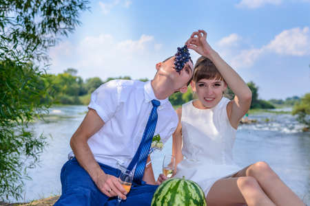 Young bridal couple sharing a meal of fresh fruit together as they sit on a blanket on the edge of a river bank celebrating after the wedding ceremonyの写真素材