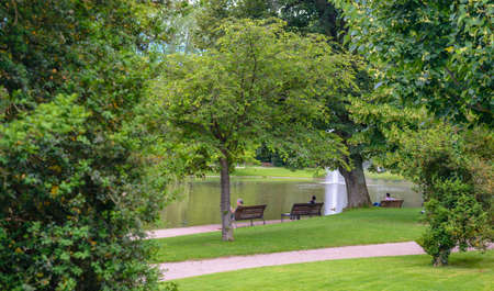 Tranquil lake in a public park with people relaxing on benches in the shade of leafy green trees overlooking the water and an ornamental fountainの写真素材