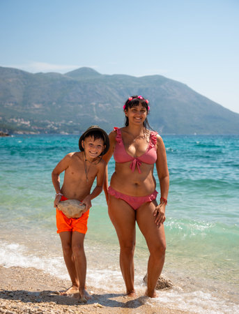Happy mother and son standing on a sunny pebble beach by the Adriatic Sea in Croatia, smiling and enjoying a warm summer day together.の写真素材