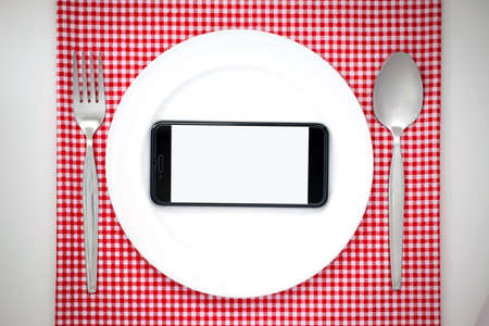 Smartphone on white dish or plate between spoon, fork on red classic checkered tablecloth texture on white table with copy space for advertise food product and otherの写真素材