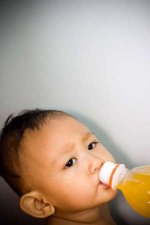 asian funny baby girl drinking orange juice from the bottle against blue wallの写真素材