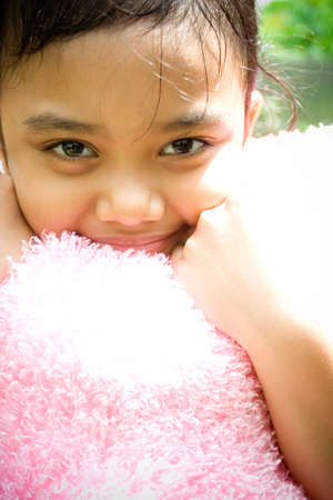 portrait of sweet asian little girl hugging a pink pillow and smiling at cameraの写真素材