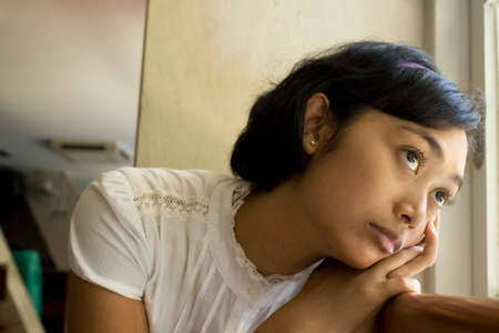 conceptual portrait of asian young woman with deep expression sitting by the window, looking out....