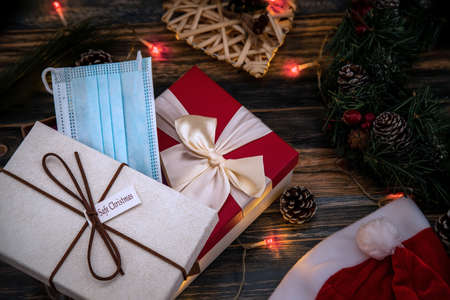 Safe COVID Christmas gifts with a medical mask over a wooden background with gift boxes, Christmas balls, branches, tree, and decorationsの写真素材
