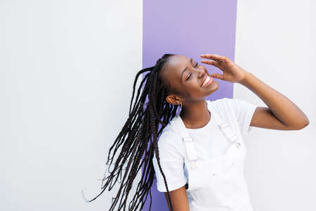Young beautiful female in white overalls having fun with closed eyes at wall
