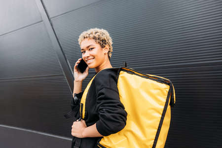 Woman courier with backpack talking on cellphone and looking at camera against the black wallの写真素材