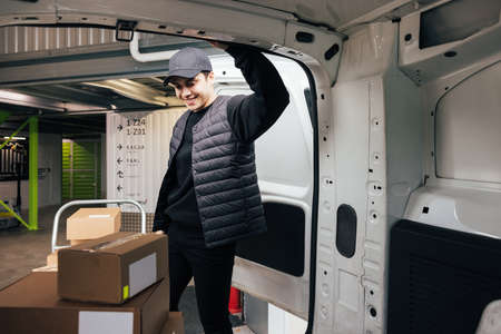 Confident courier leaning on van looking on cardboard boxes