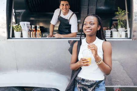 Smiling female customer holding orange juice against food truck with the ownerの写真素材