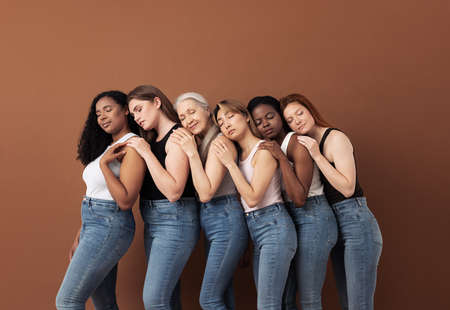 Diverse women in casuals embracing each other while standing with closed eyes over brown backgroundの写真素材
