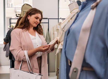 Woman standing in a small fashion boutique and looking at fabric of a shirt on a mannequin