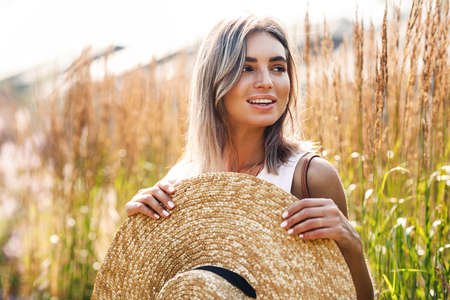 Beautiful smiling woman holding straw hat in hands standing outdoorsの写真素材