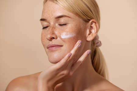 Close up of beautiful young female with cream on a face. Smiling woman with closed eyes touching cheek with fingers.の写真素材