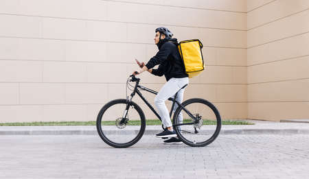 Side view of a young messenger with termal backpack man riding his bike and using mobile phone in the cityの写真素材