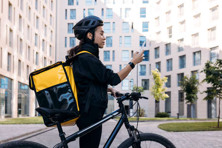 Side view of delivery woman with backpack looking at smartphone while standing in the city against apartment buildingの写真素材