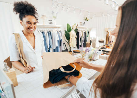 Unrecognizable clothing store owner packing the clothes in a paper bag. Smiling buyer looking at...