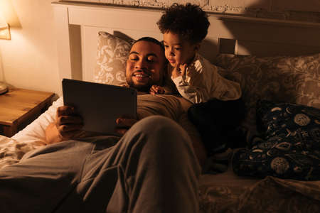 Smiling dad and his little son in bed watching content on a digital tabletの写真素材