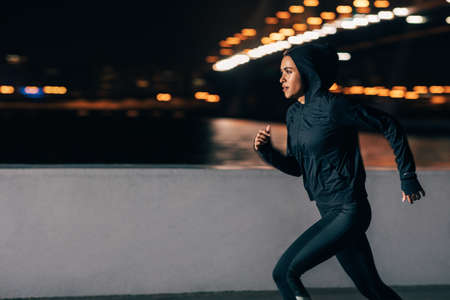 Side view of a woman in hoodie running at night. Middle east female jogger exercising outdoors.の写真素材