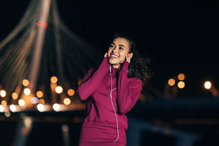 Cheerful woman in sport clothes enjoying music while standing at nightの写真素材