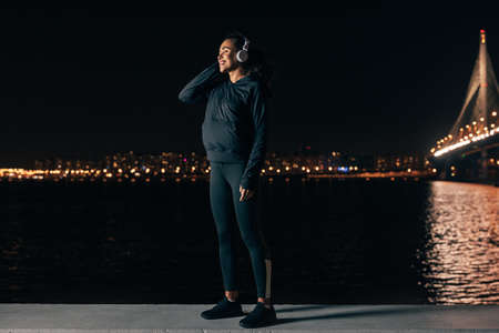 Young female in sport clothes standing on embankment listening to music against night city lights