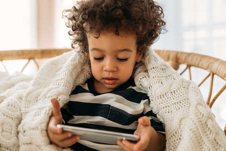 Little cute boy with curly hair sitting on an armchair under a plaid and holding a smartphone
