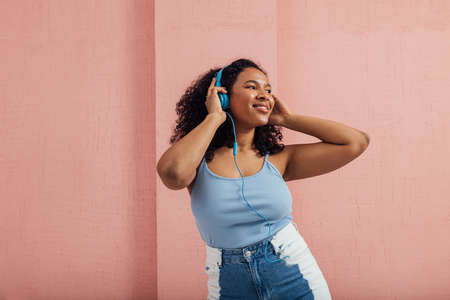 Smiling woman in casuals standing against pink wall listening to music and looking awayの写真素材