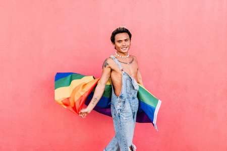 Smiling young gay man standing with pride flag. Happy guy in casuals holding LGBT flag outdoors.の写真素材