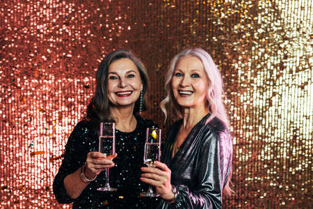 Stylish senior females standing together against glitter backdrop holding glasses of champagne