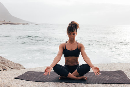 Woman in lotus pose sitting on yoga mat by the ocean at sunsetの写真素材