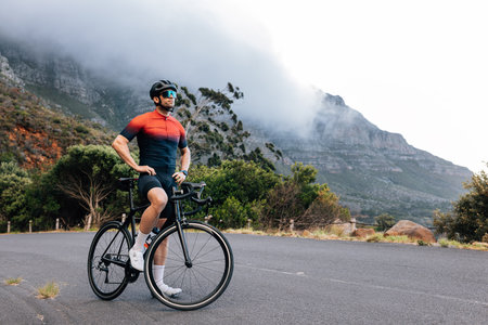 Young male cyclist relaxing after training with hands on a hips. Professional athlete with road bike enjoying the view while standing on a road in wild terrain.の写真素材