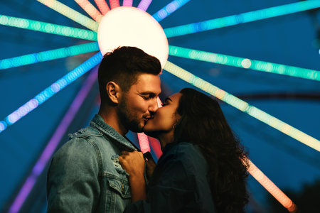 Young couple kissing in front of a Ferris wheel. Girl kissing her boyfriend in an amusement park at...