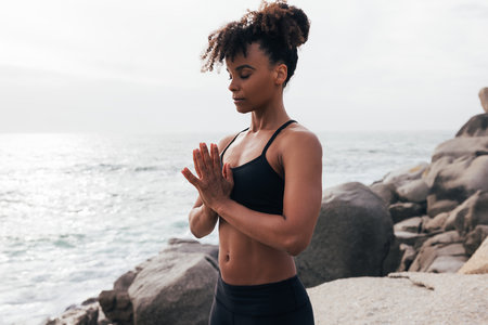 Young woman with folded hands standing at sunset by ocean. Female meditating outdoors with closed eyes.の写真素材