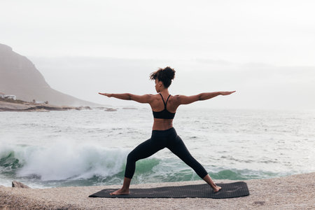Back view of female practicing yoga in warrior pose by the ocean at bayの写真素材