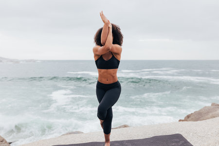 Woman standing in Eagle yoga pose against an ocean at sunsetの写真素材