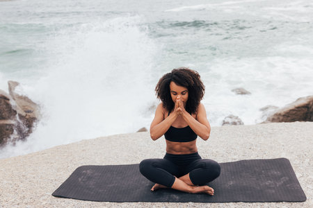 Young woman sitting on the beach meditating with eyes closed. Female with hands together and waves behind doing yoga.の写真素材