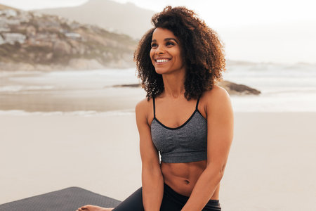 Portrait of a beautiful young woman with curly hair wearing sportswear sitting on beach relaxing at...