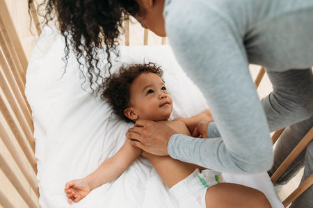 Woman leaning into a crib. Young mother looking at her son and holding hands.