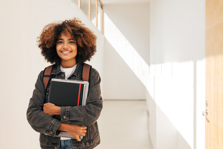 Portrait of a smiling student holding notebook and laptop while standing in corridorの写真素材