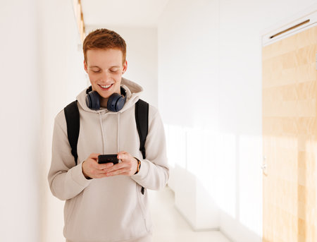 Smiling guy using smartphone while walking in college corridorの写真素材
