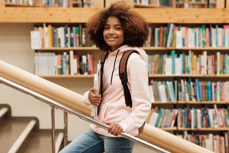 Beautiful girl with curly hair standing in library against bbokshelves and looking at cameraの写真素材
