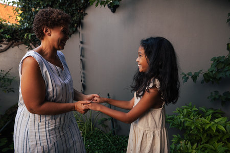 Granny and granddaughter standing together in the backyard holding hands and looking at each other. Kid and grandma spending time together.の写真素材