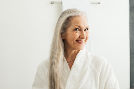 Smiling woman with long white hair at morning in bathroom. Aged female in bathrobe looking at her reflection in the mirror.の写真素材