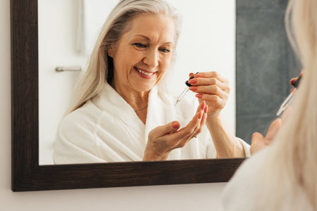 Reflection in mirror of a smiling aged woman with a pipette. Smiling senior female in a bathroom using liquid face treatment.の写真素材
