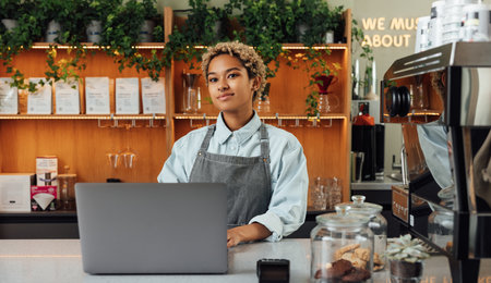 Young female coffee shop owner at counter with a laptop. Woman barista in an apron working in a coffee shop.の写真素材