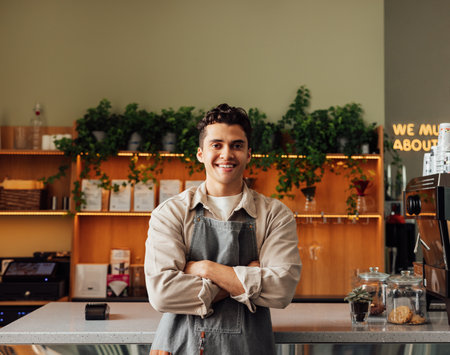 Coffee shop owner in an apron standing with crossed arms and smiling. Young handsome man leaning counter looking at a camera in a coffee shop.の写真素材
