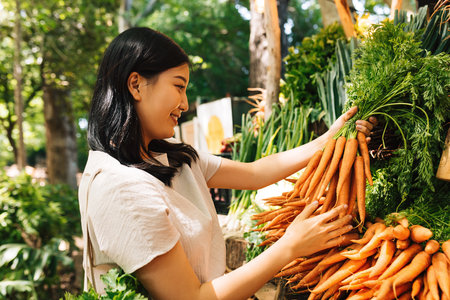 Smiling buyer holding a bunch of carrots. Asian woman choosing vegetables in the local market.の写真素材
