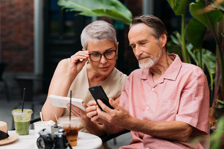 Aged woman looking at her husband's smartphone in an outdoor cafe. Senior man showing his wife something on his smartphone.の写真素材