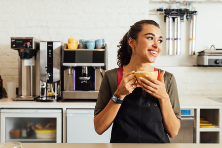 Female waitress holding coffee mug while standing at table in coffee shopの写真素材
